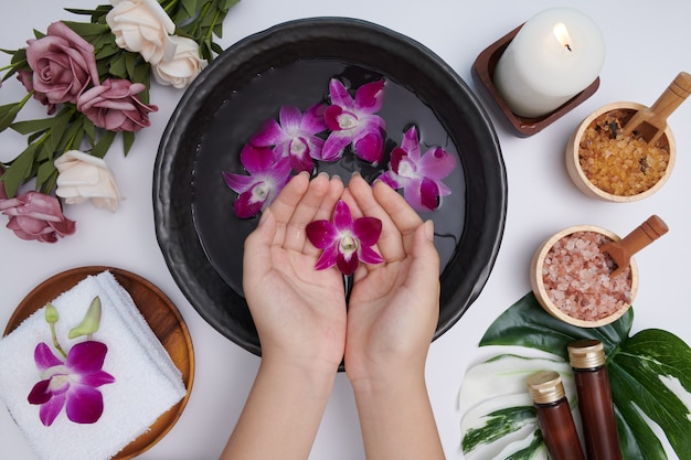 Woman soaking her hands in bowl of water and flowers, spa treatment and product for female feet and hand spa, massage pebble, perfumed flowers water and candles, relaxation. flat lay. top view. Free Photo