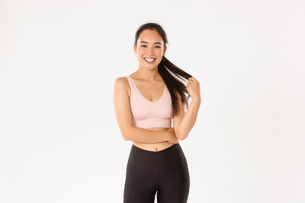 Sport, wellbeing and active lifestyle concept. smiling pretty asian girl in sportswear, playing with ponytail and looking happy at camera after good gym workout, standing white background.