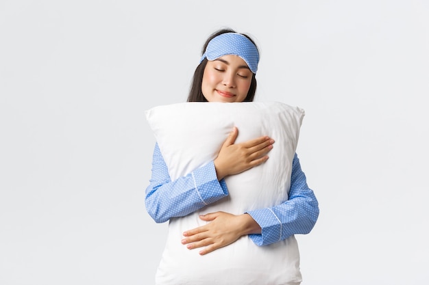 Free photo beautiful smiling asian girl in sleeping mask and pajama, having sweet dream, hugging pillow with silly grin and closed eyes, standing over white background, unwilling wake-up.