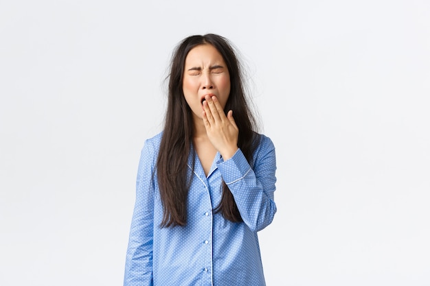 Free photo tired yawning asian girl in blue pajamas, yawning sleepy with messy haircut after waking up, cover opened mouth with hand, standing in jammies after rough night, white background.