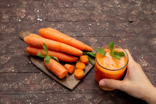 Free photo front view fresh carrot juice inside long glass with leaf and along with fresh carrots on the brown desk